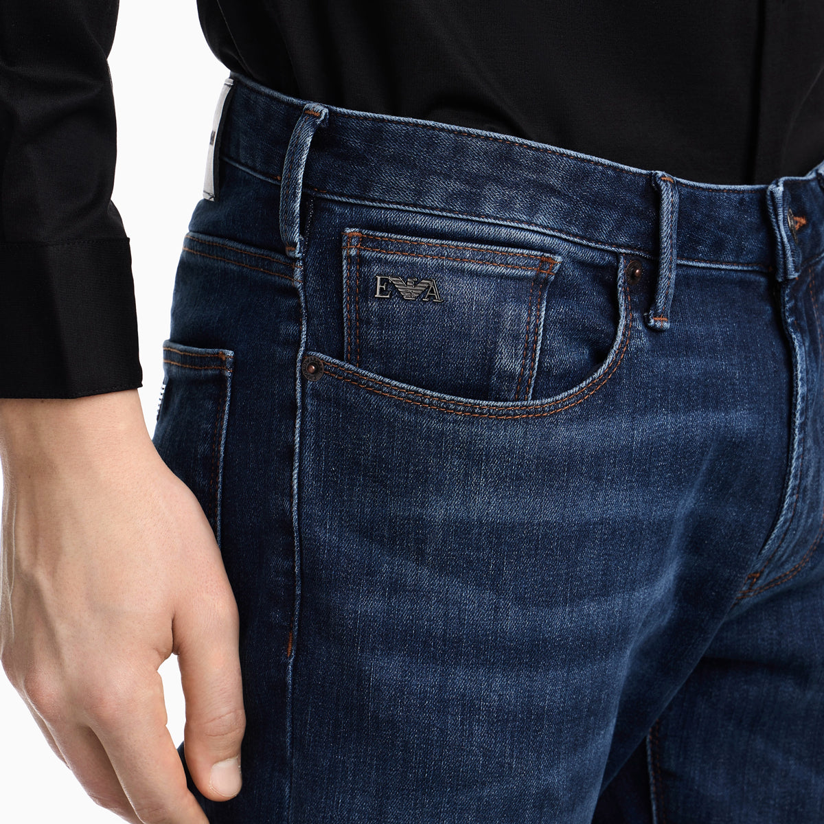 Close-up of a person wearing dark blue denim jeans with a black long-sleeve shirt. The jeans feature a visible front pocket with a small embroidered logo and contrast stitching. Emporio Armani Jeans available at Sakks Menswear in Southampton.