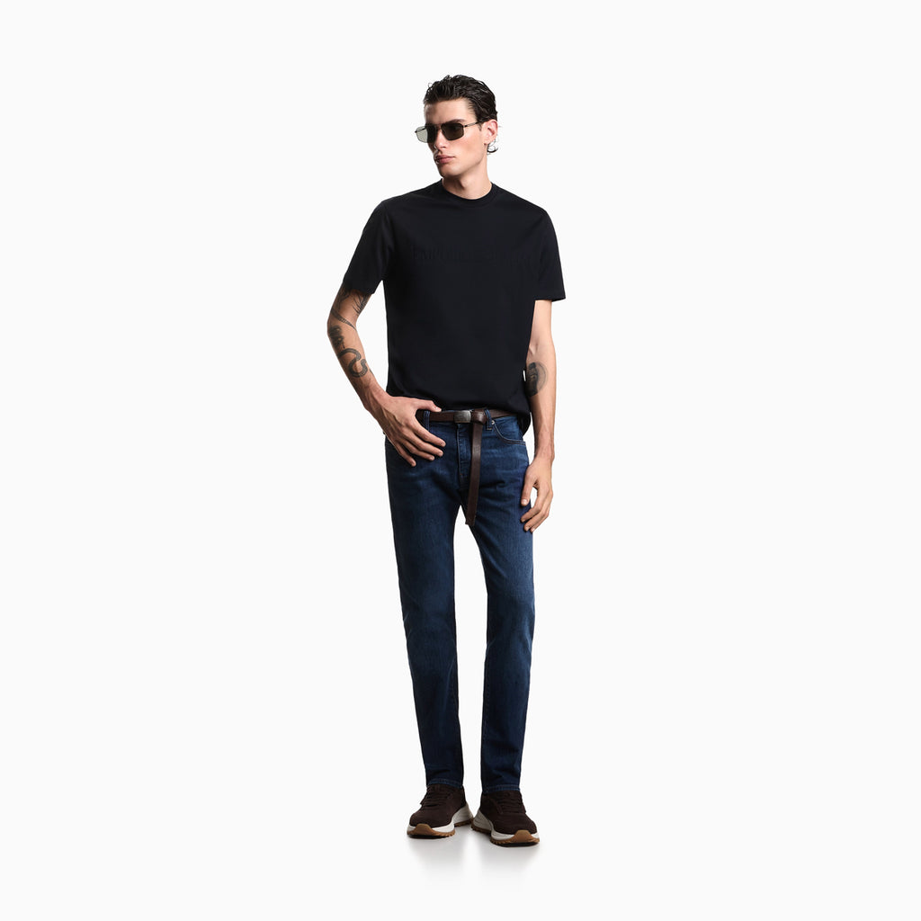 A man modeling a black short-sleeve crewneck t-shirt and dark blue slim-fit jeans with a brown leather belt, styled with sunglasses, a black watch, and brown sneakers, set against a plain white studio background. Emporio Armani Jeans available at Sakks Menswear in Southampton