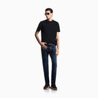 A man modeling a black short-sleeve crewneck t-shirt and dark blue slim-fit jeans with a brown leather belt, styled with sunglasses, a black watch, and brown sneakers, set against a plain white studio background. Emporio Armani Jeans available at Sakks Menswear in Southampton