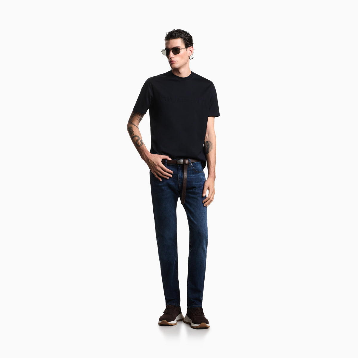 A man modeling a black short-sleeve crewneck t-shirt and dark blue slim-fit jeans with a brown leather belt, styled with sunglasses, a black watch, and brown sneakers, set against a plain white studio background. Emporio Armani Jeans available at Sakks Menswear in Southampton