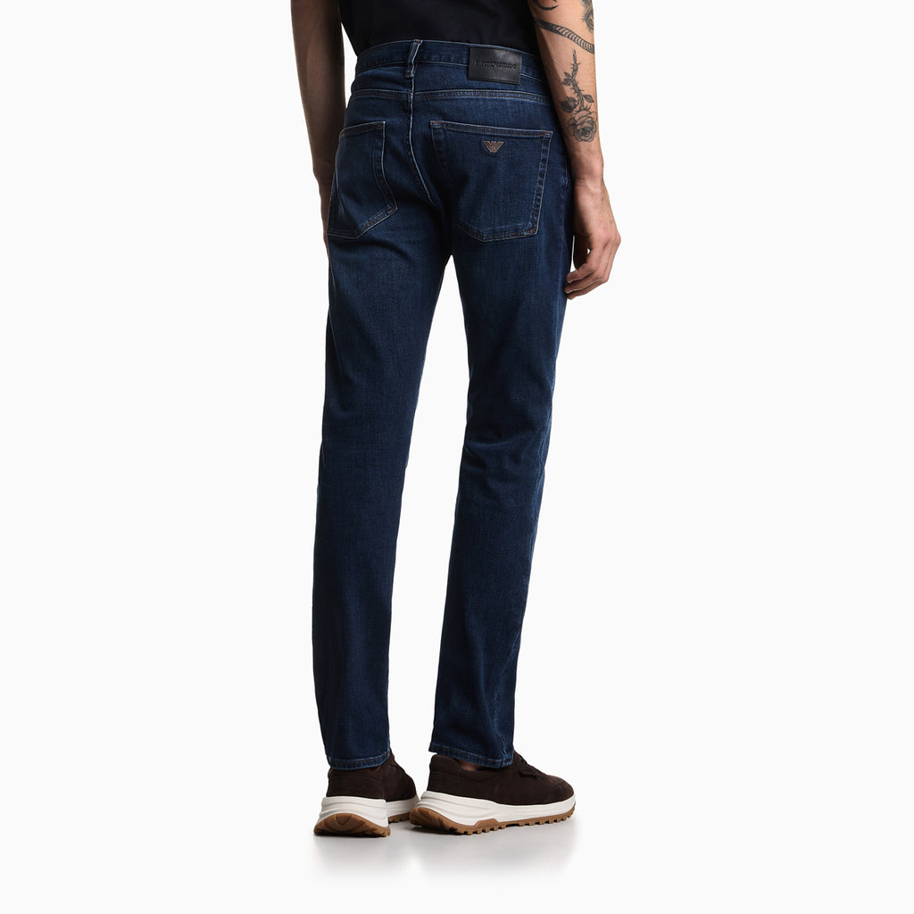 Rear view of a person wearing dark blue slim-fit jeans with brown sneakers, shown against a plain white studio background. Emporio Armani Jeans available at Sakks Menswear in Southampton