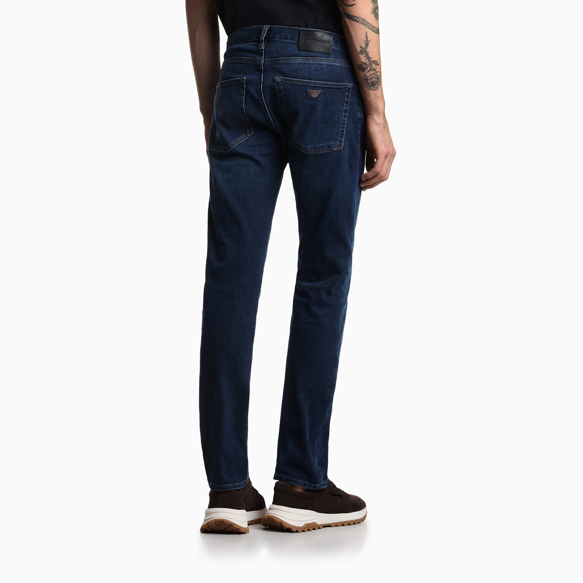 Rear view of a person wearing dark blue slim-fit jeans with brown sneakers, shown against a plain white studio background. Emporio Armani Jeans available at Sakks Menswear in Southampton