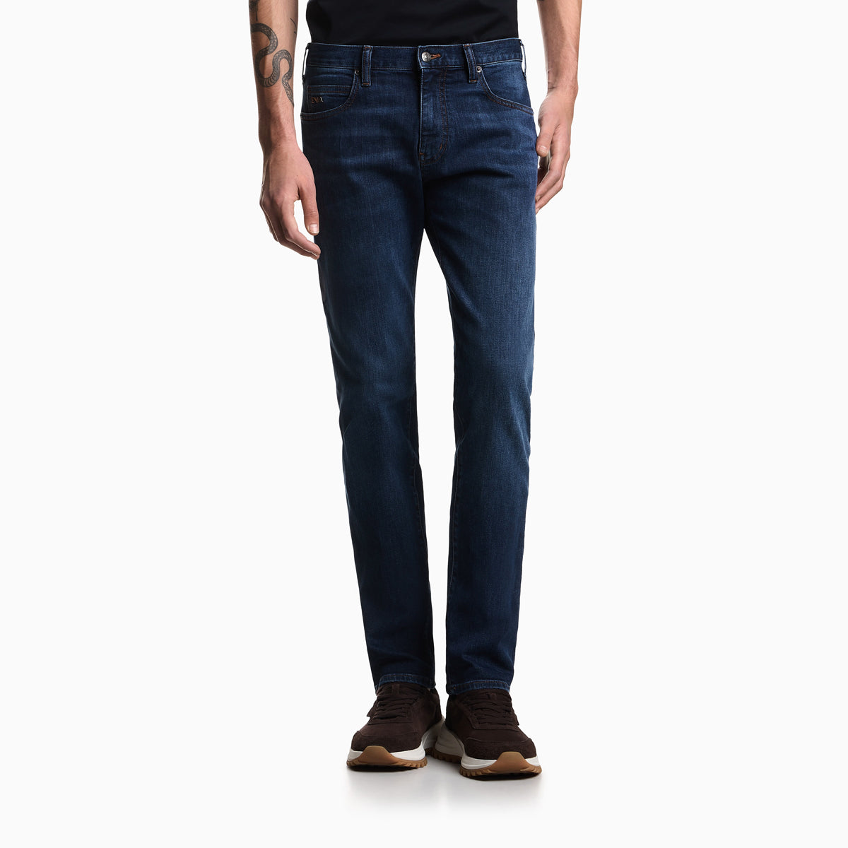 A person wearing dark blue slim-fit jeans, a black t-shirt, and brown sneakers, shown from the waist down against a plain white studio background. Emporio Armani Jeans available at Sakks Menswear in Southampton