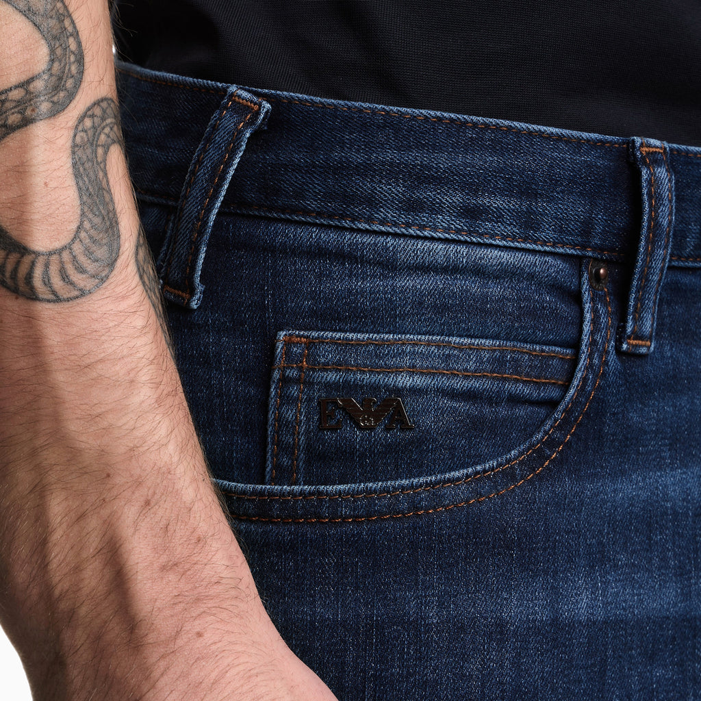 Close-up of a person wearing dark blue denim jeans, showing the back pocket with an embossed logo and the side of the model's tattooed arm. Emporio Armani Jeans available at Sakks Menswear in Southampton