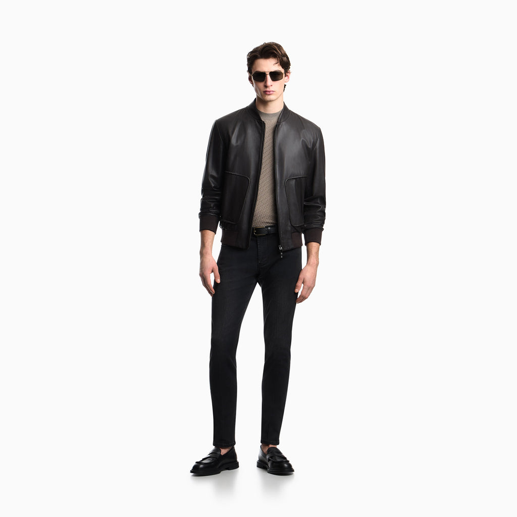A male model wearing a black leather bomber jacket, black slim-fit jeans, a beige ribbed turtleneck, and black leather loafers, standing against a solid white background. Emporio Armani Jeans available at Sakks Menswear in Southampton.