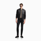 A male model wearing a black leather bomber jacket, black slim-fit jeans, a beige ribbed turtleneck, and black leather loafers, standing against a solid white background. Emporio Armani Jeans available at Sakks Menswear in Southampton.