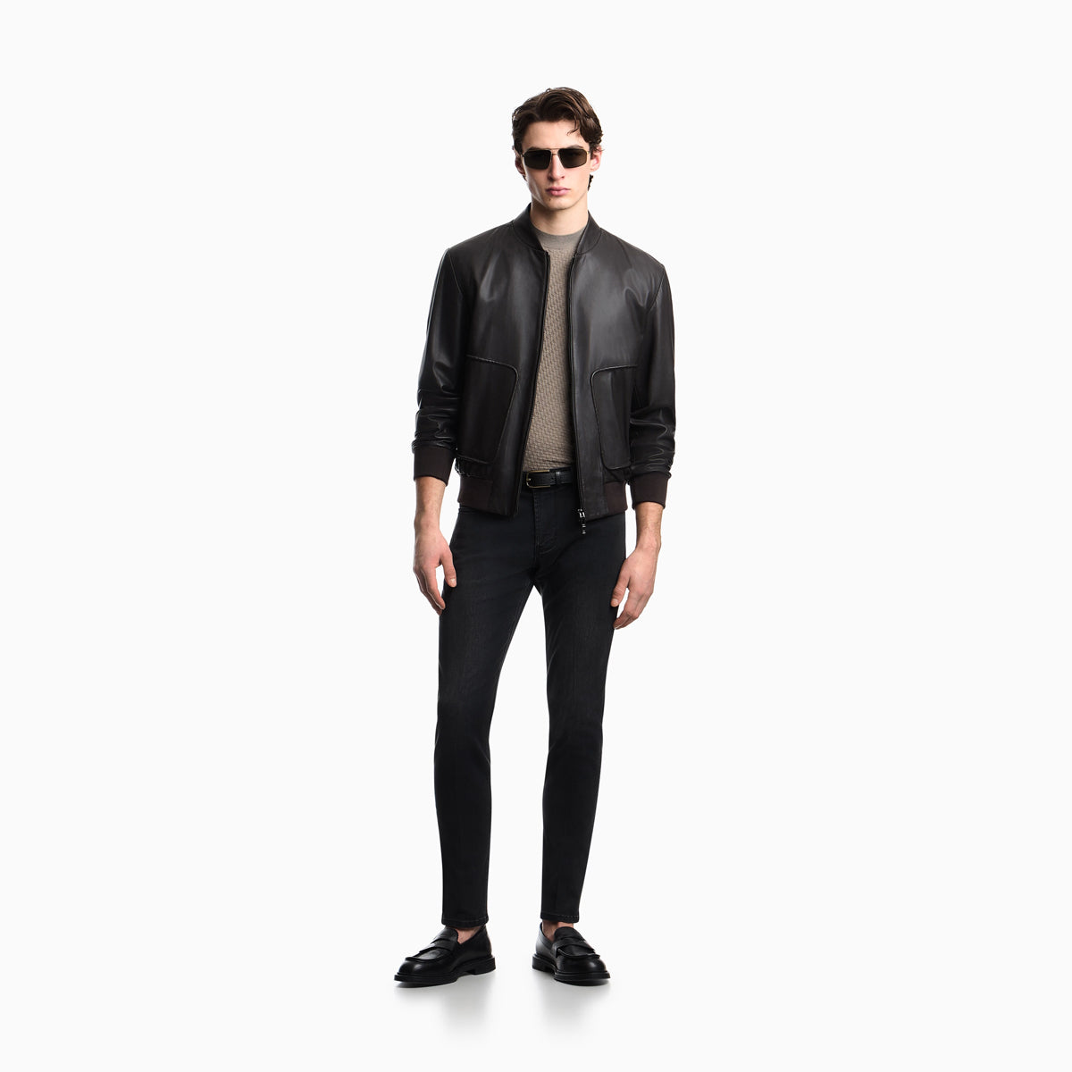 A male model wearing a black leather bomber jacket, black slim-fit jeans, a beige ribbed turtleneck, and black leather loafers, standing against a solid white background. Emporio Armani Jeans available at Sakks Menswear in Southampton.