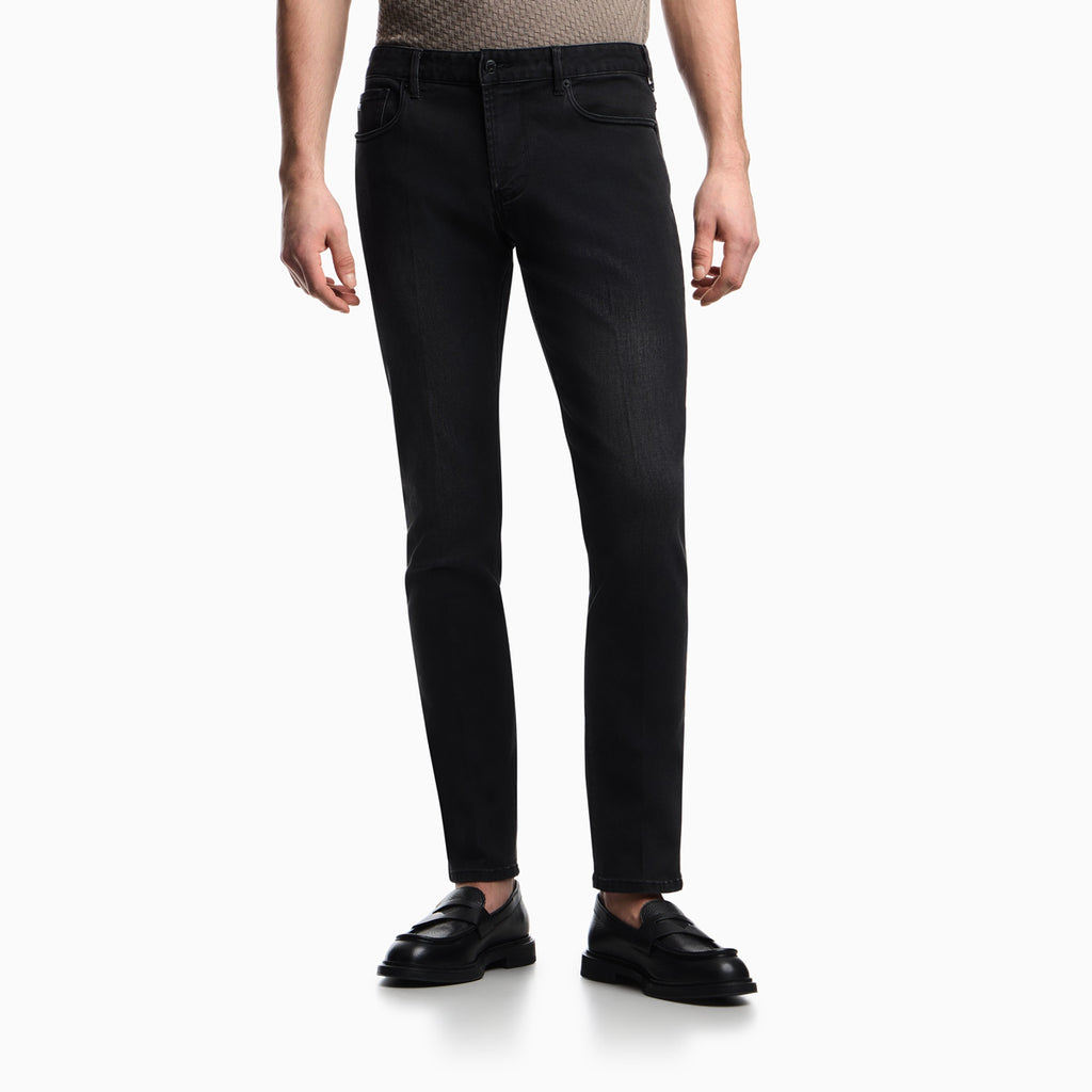 A model wearing slim-fit black jeans, a beige patterned knit top, and black leather loafers, shown from the waist down on a clean white studio background. Emporio Armani Jeans available at Sakks Menswear in Southampton.