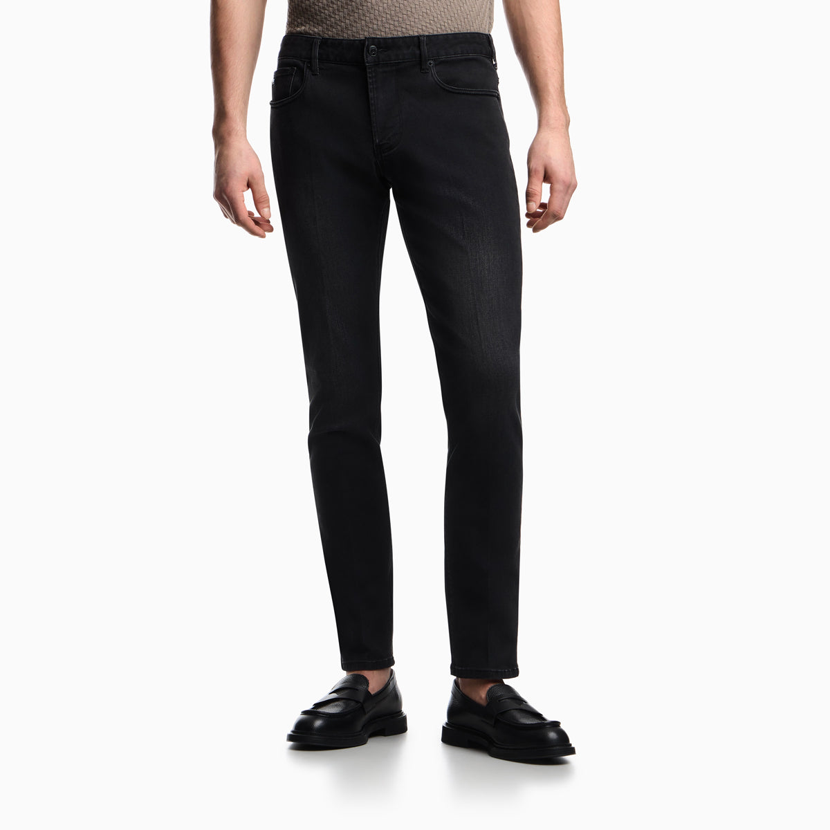 A model wearing slim-fit black jeans, a beige patterned knit top, and black leather loafers, shown from the waist down on a clean white studio background. Emporio Armani Jeans available at Sakks Menswear in Southampton.