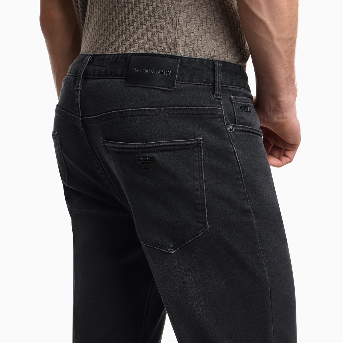 Close-up back view of a person wearing dark charcoal gray Emporio Armani jeans, showing the back pocket, belt loop, and brand patch on a light gray background. Emporio Armani Jeans available at Sakks Menswear in Southampton.
