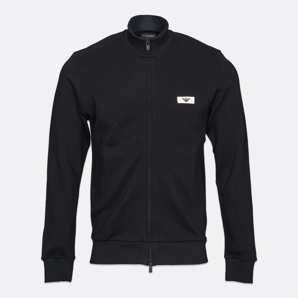 A black zip-up track jacket with a stand collar, featuring a small white rectangular chest logo with an eagle emblem, displayed on a light gray background. Emporio Armani Sweatshirt available at Sakks Menswear in Southampton.