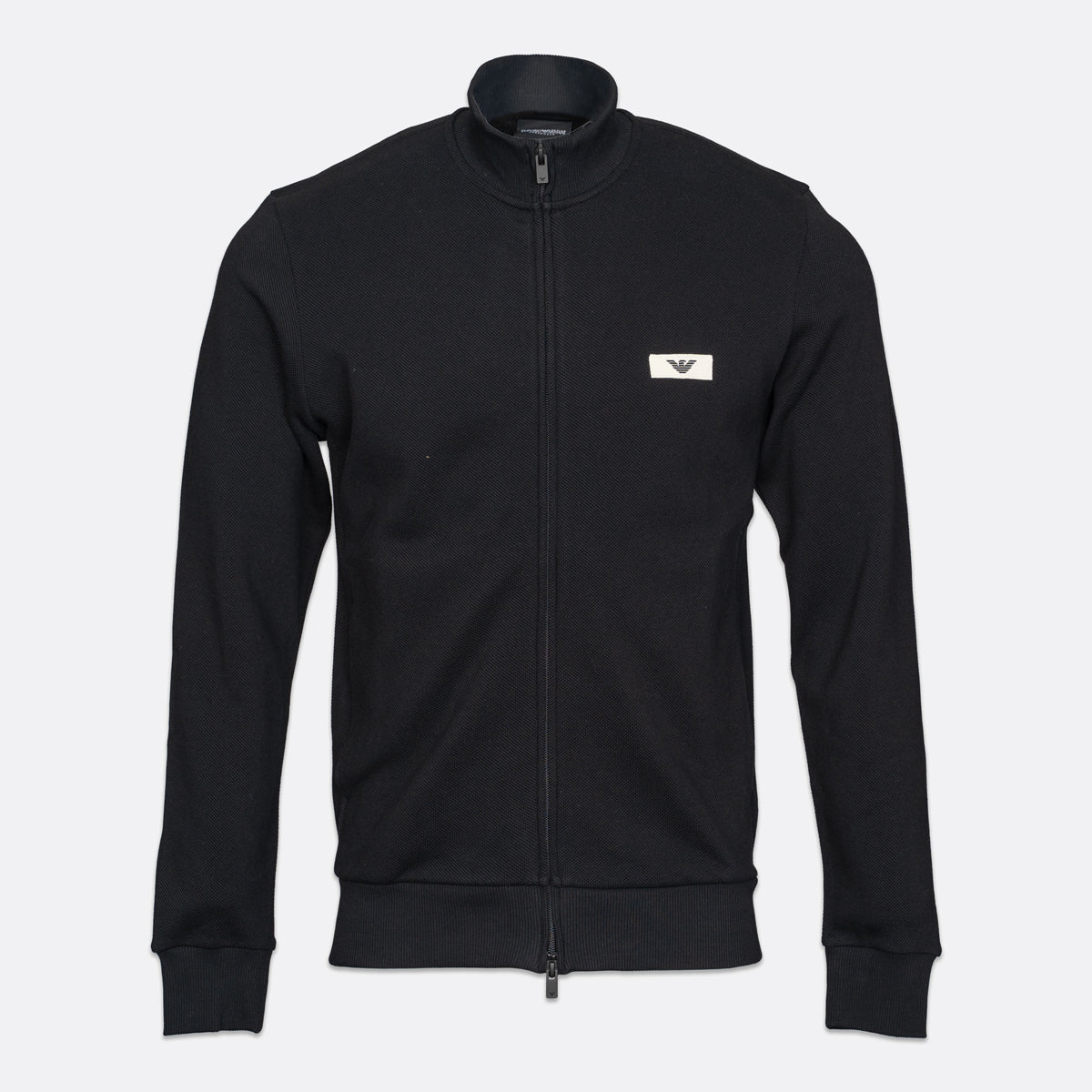 A black zip-up track jacket with a stand collar, featuring a small white rectangular chest logo with an eagle emblem, displayed on a light gray background. Emporio Armani Sweatshirt available at Sakks Menswear in Southampton.