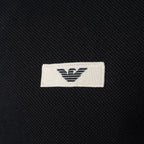 Close-up of a black textured knit garment featuring a white rectangular woven label with the black embroidered Emporio Armani eagle logo. Emporio Armani Sweatshirt available at Sakks Menswear in Southampton.