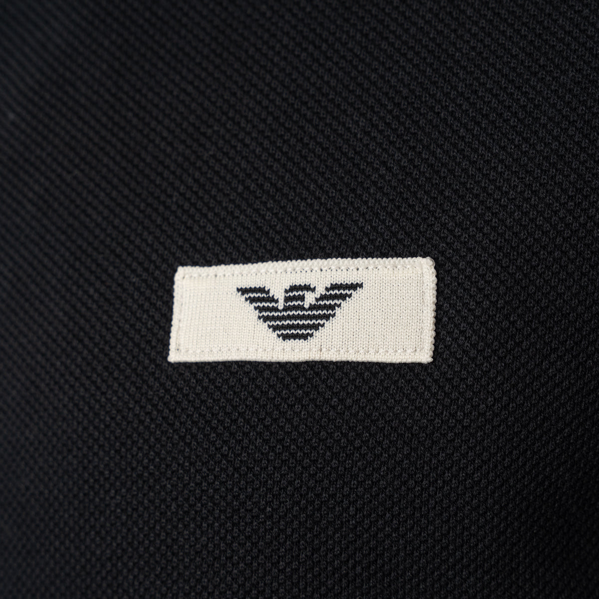Close-up of a black textured knit garment featuring a white rectangular woven label with the black embroidered Emporio Armani eagle logo. Emporio Armani Sweatshirt available at Sakks Menswear in Southampton.