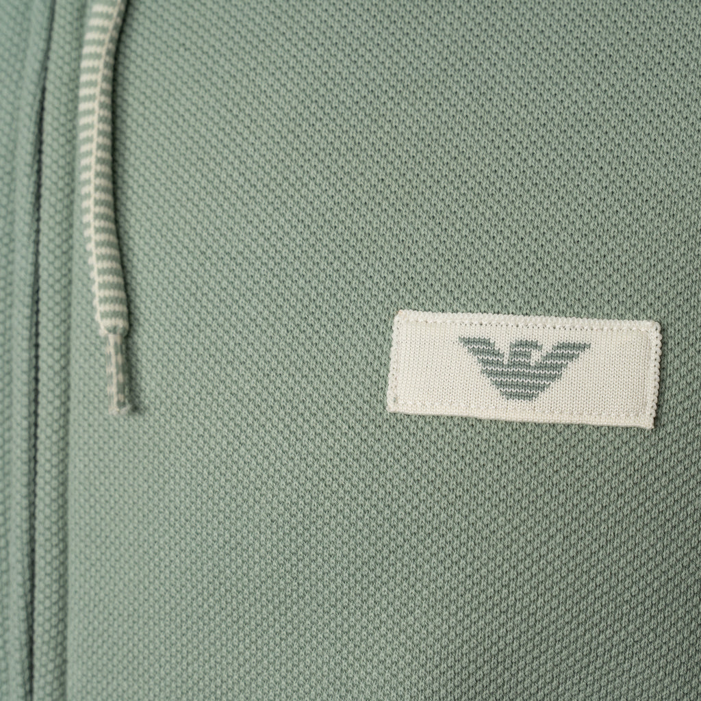 Close-up of a sage green textured fabric item featuring a white rectangular patch with the grey embroidered Emporio Armani eagle logo. Emporio Armani Hoodie available at Sakks Menswear in Southampton.