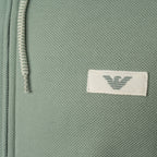 Close-up of a sage green textured fabric item featuring a white rectangular patch with the grey embroidered Emporio Armani eagle logo. Emporio Armani Hoodie available at Sakks Menswear in Southampton.