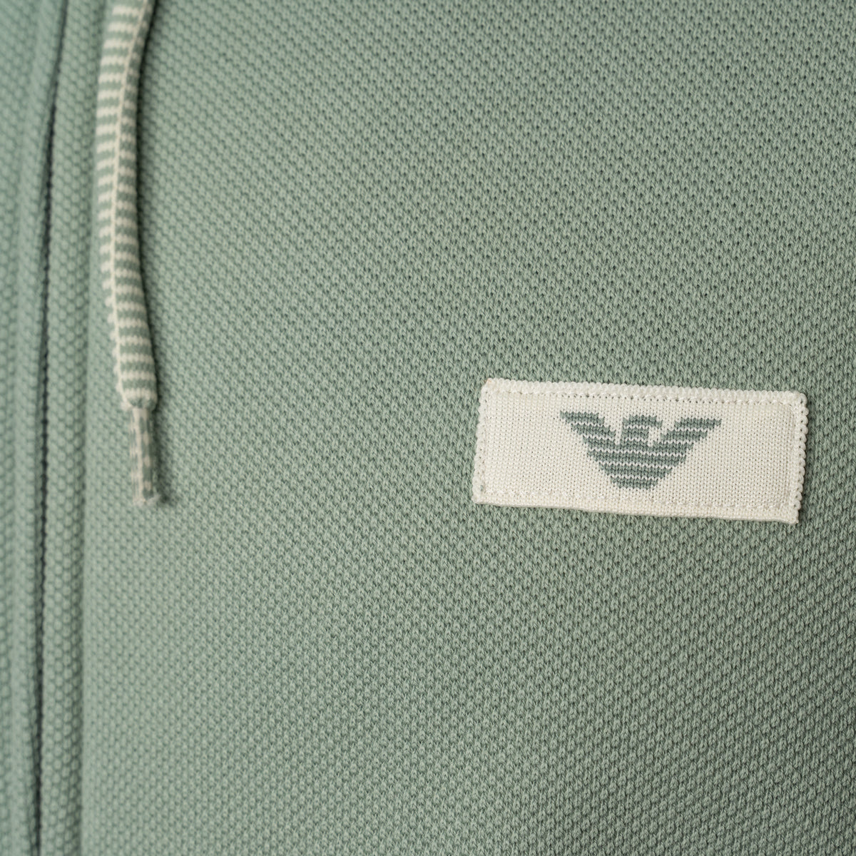 Close-up of a sage green textured fabric item featuring a white rectangular patch with the grey embroidered Emporio Armani eagle logo. Emporio Armani Hoodie available at Sakks Menswear in Southampton.