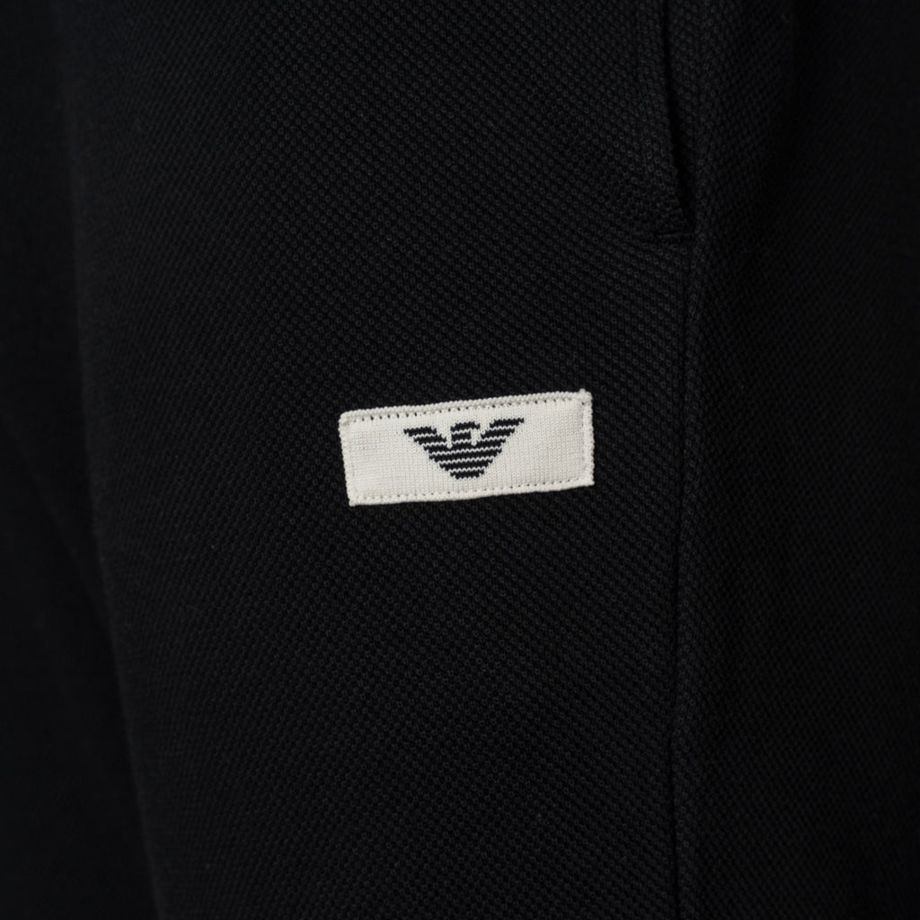 Close-up of a black textured fabric garment featuring a small white rectangular patch with the black embroidered Emporio Armani eagle logo. Emporio Armani Joggers available at Sakks Menswear in Southampton.