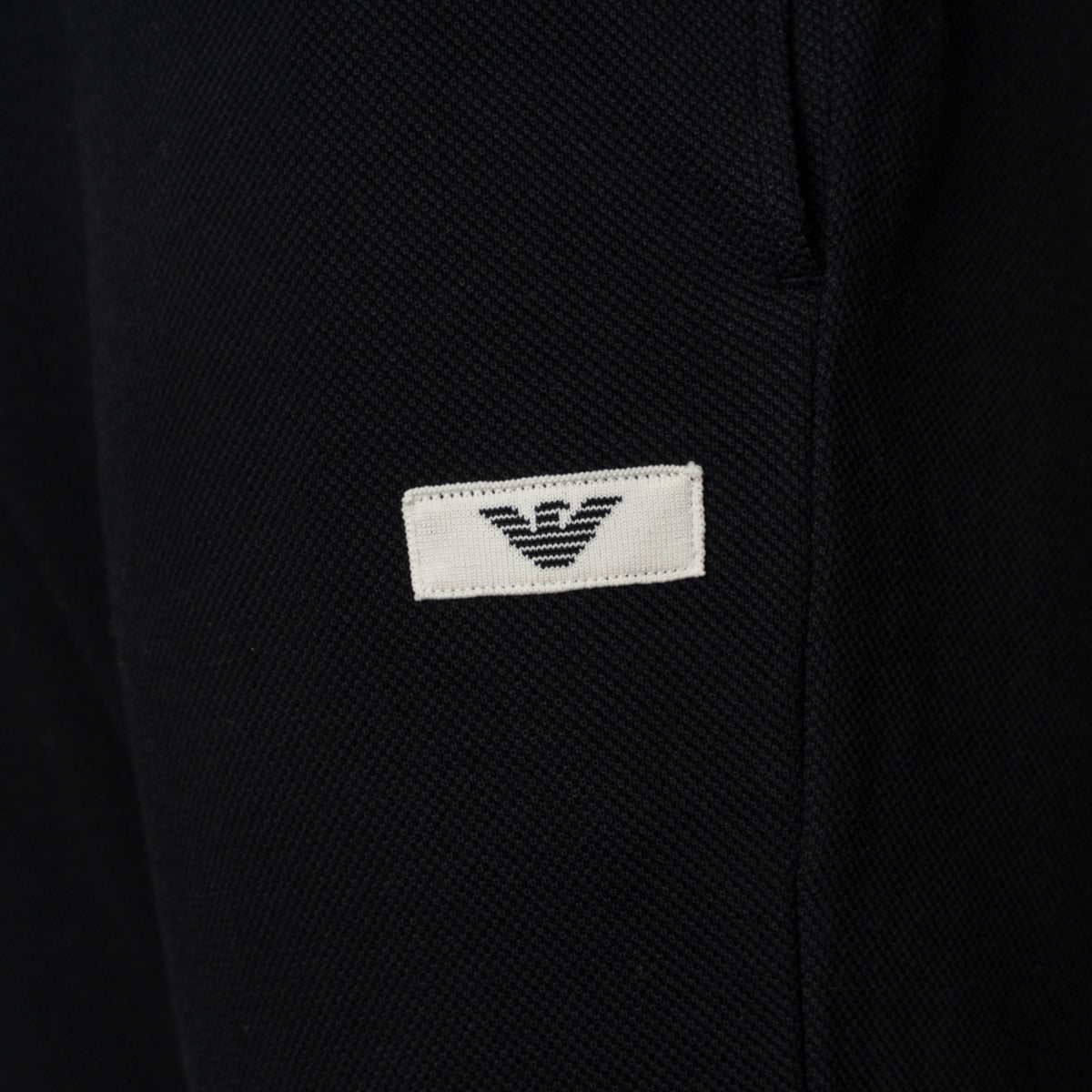 Close-up of a black textured fabric garment featuring a small white rectangular patch with the black embroidered Emporio Armani eagle logo. Emporio Armani Joggers available at Sakks Menswear in Southampton.