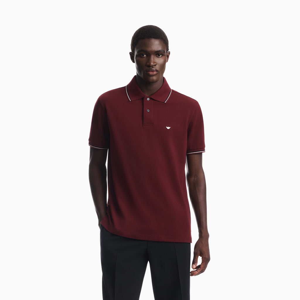 A man wearing a short-sleeve polo shirt in burgundy with white piping on the collar and sleeve cuffs, paired with dark trousers, against a plain white studio background. Emporio Armani Polo available at Sakks Menswear in Southampton