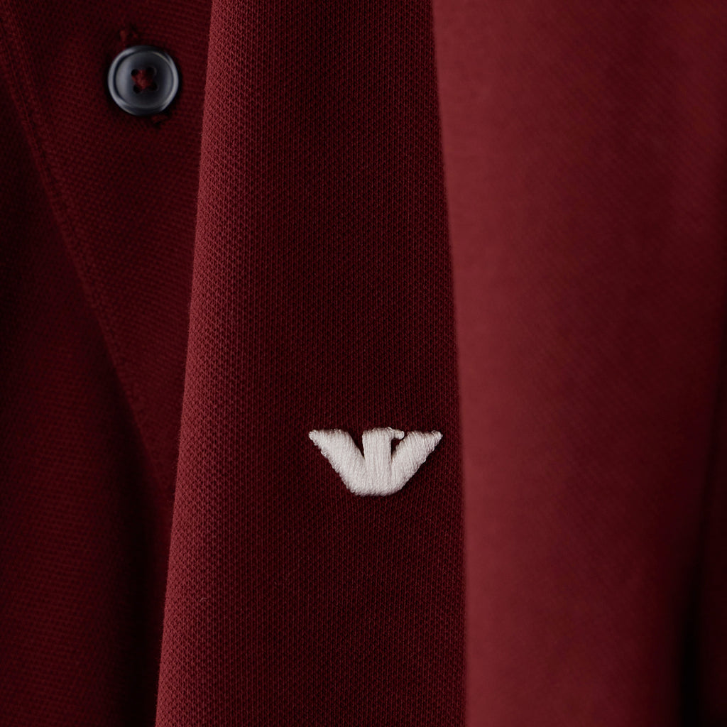 Close-up of a dark red textured knit garment with a small white embroidered logo and a dark button, showing fabric texture and branding. Emporio Armani Polo available at Sakks Menswear in Southampton