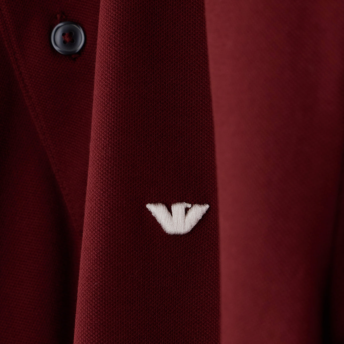 Close-up of a dark red textured knit garment with a small white embroidered logo and a dark button, showing fabric texture and branding. Emporio Armani Polo available at Sakks Menswear in Southampton