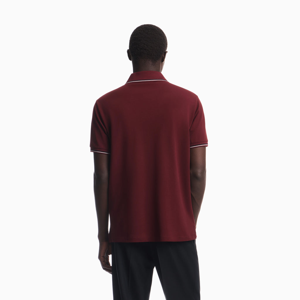 A man viewed from behind wearing a short-sleeve polo shirt in burgundy with thin white piping on the collar and sleeve cuffs, paired with dark trousers, against a plain white studio background. Emporio Armani Polo available at Sakks Menswear in Southampton