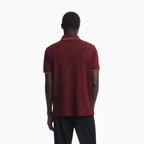 A man viewed from behind wearing a short-sleeve polo shirt in burgundy with thin white piping on the collar and sleeve cuffs, paired with dark trousers, against a plain white studio background. Emporio Armani Polo available at Sakks Menswear in Southampton