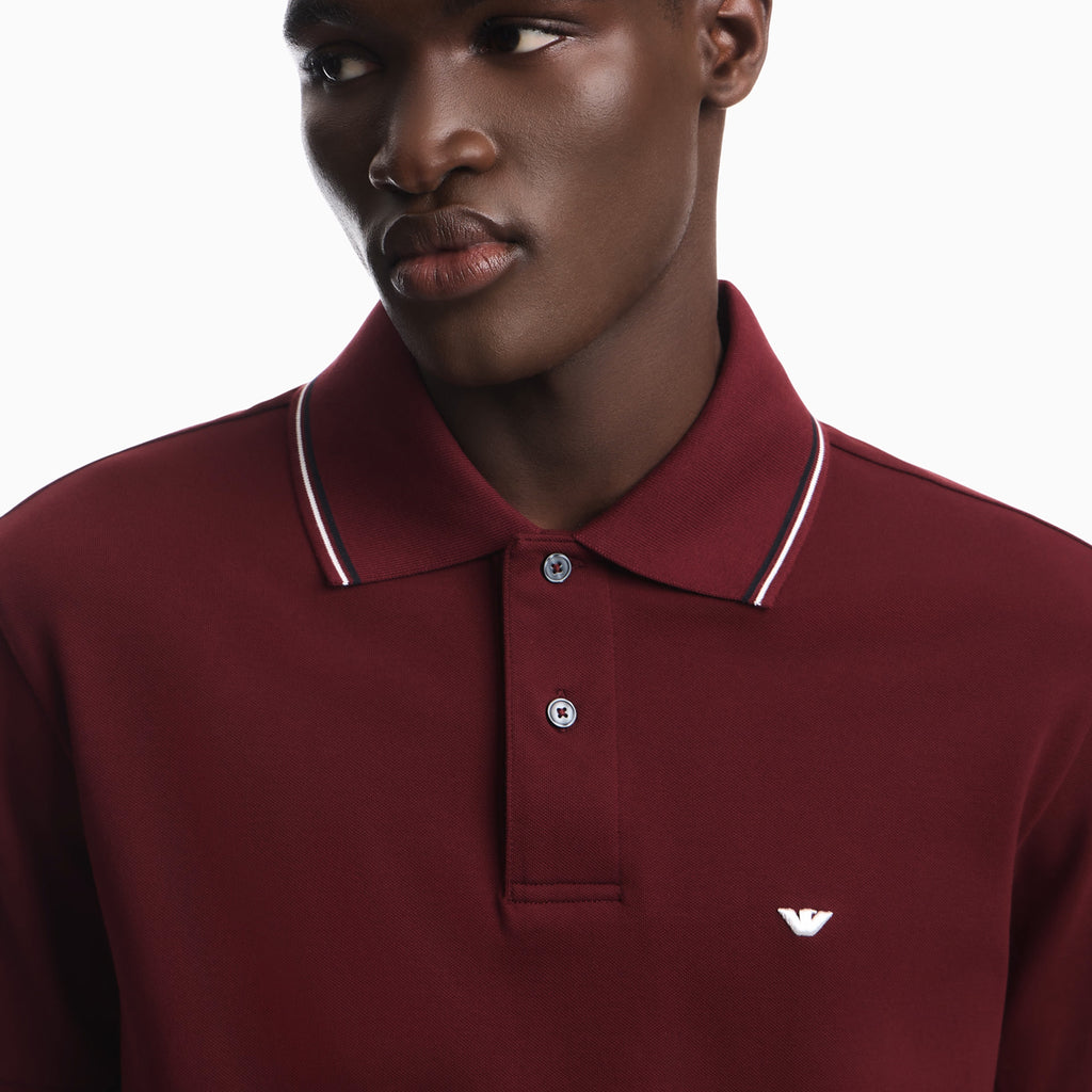 A close-up of a man wearing a maroon polo shirt with a white and navy striped collar and a small white embroidered eagle logo on the chest. Emporio Armani Polo available at Sakks Menswear in Southampton
