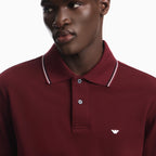 A close-up of a man wearing a maroon polo shirt with a white and navy striped collar and a small white embroidered eagle logo on the chest. Emporio Armani Polo available at Sakks Menswear in Southampton