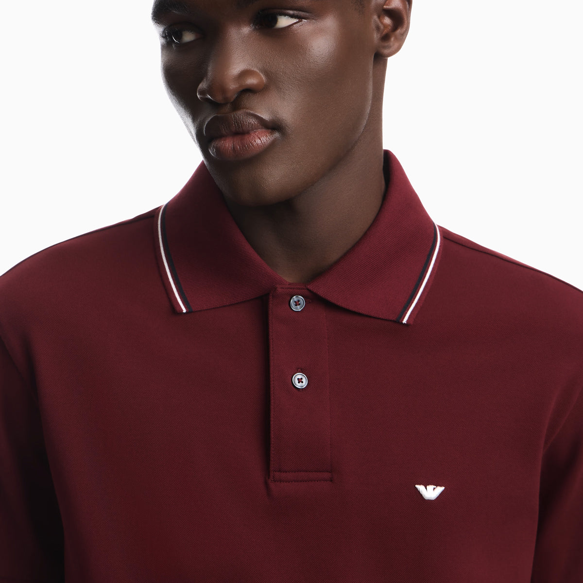 A close-up of a man wearing a maroon polo shirt with a white and navy striped collar and a small white embroidered eagle logo on the chest. Emporio Armani Polo available at Sakks Menswear in Southampton
