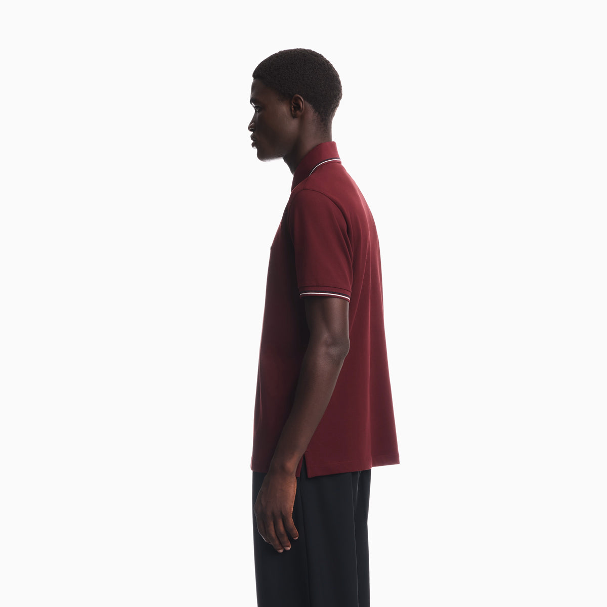 A man in profile wearing a short-sleeve polo shirt in deep burgundy with thin white trim on the collar and sleeve cuffs, paired with dark trousers, against a plain white studio background. Emporio Armani Polo available at Sakks Menswear in Southampton