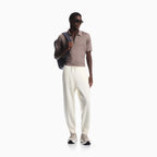 A man modeling a taupe short-sleeve polo shirt, off-white drawstring sweatpants, and beige sneakers, carrying a dark blue backpack against a plain white background. Emporio Armani olo available at Sakks Menswear in Southampton.