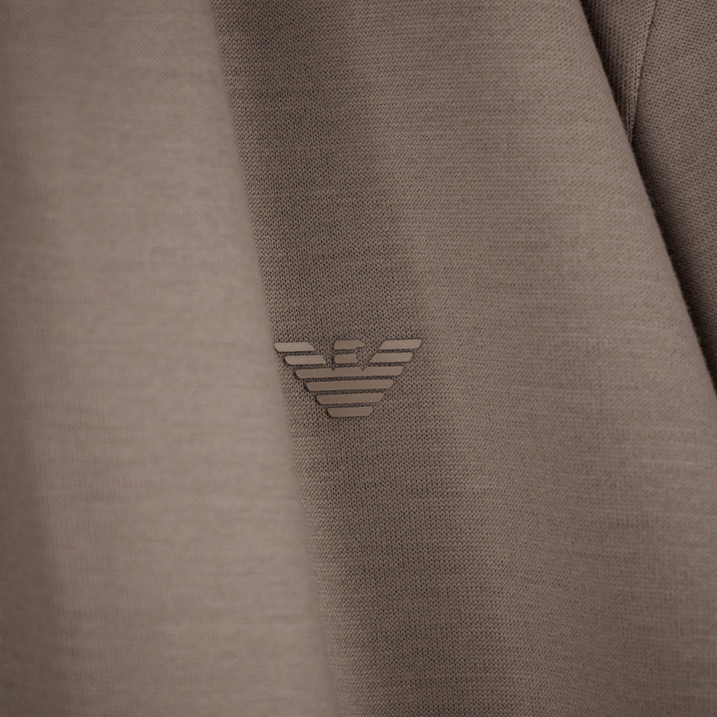 Close-up of a taupe-brown textured fabric with a small metallic silver embroidered logo in the center. Emporio Armani olo available at Sakks Menswear in Southampton.