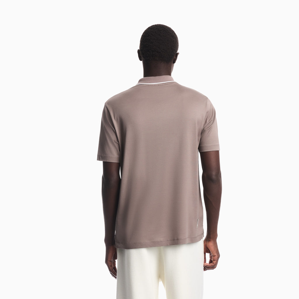 A man viewed from the back wearing a short-sleeve polo shirt in a muted taupe-brown color with a thin white stripe detail at the collar, paired with off-white trousers, against a plain white background. Emporio Armani olo available at Sakks Menswear in Southampton.