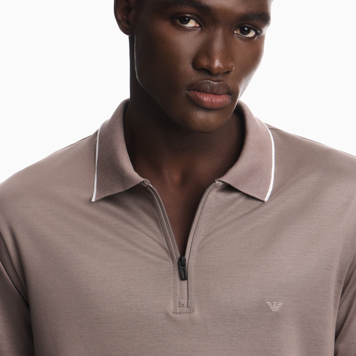 A close-up of a man wearing a taupe-brown half-zip polo shirt with white piping on the collar and a small embroidered eagle logo on the chest, set against a plain white background. Emporio Armani olo available at Sakks Menswear in Southampton.