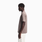 A man in profile wearing a short-sleeve polo shirt in a muted taupe-beige color with white pants, photographed against a clean white studio background. Emporio Armani olo available at Sakks Menswear in Southampton.