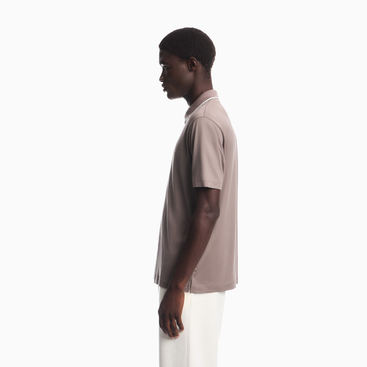 A man in profile wearing a short-sleeve polo shirt in a muted taupe-beige color with white pants, photographed against a clean white studio background. Emporio Armani olo available at Sakks Menswear in Southampton.