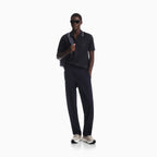A man modeling a dark navy polo shirt with white piping, matching dark navy trousers, and light beige sneakers, standing against a solid white background. Emporio Armani Polo available at Sakks Menswear in Southampton.