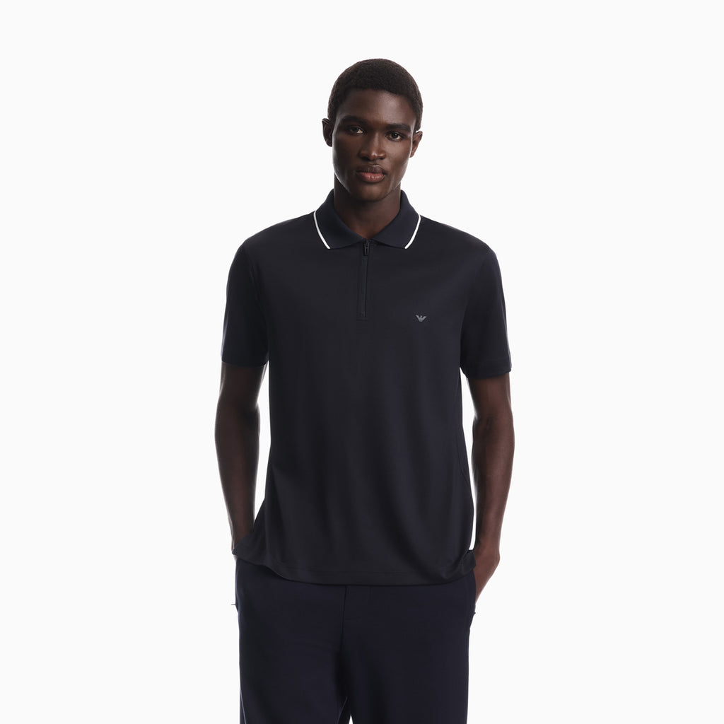 A man wearing a dark navy blue polo shirt with white piping on the collar and a small embroidered eagle logo on the chest, paired with matching dark trousers, set against a plain white background. Emporio Armani Polo available at Sakks Menswear in Southampton.