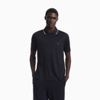 A man wearing a dark navy blue polo shirt with white piping on the collar and a small embroidered eagle logo on the chest, paired with matching dark trousers, set against a plain white background. Emporio Armani Polo available at Sakks Menswear in Southampton.