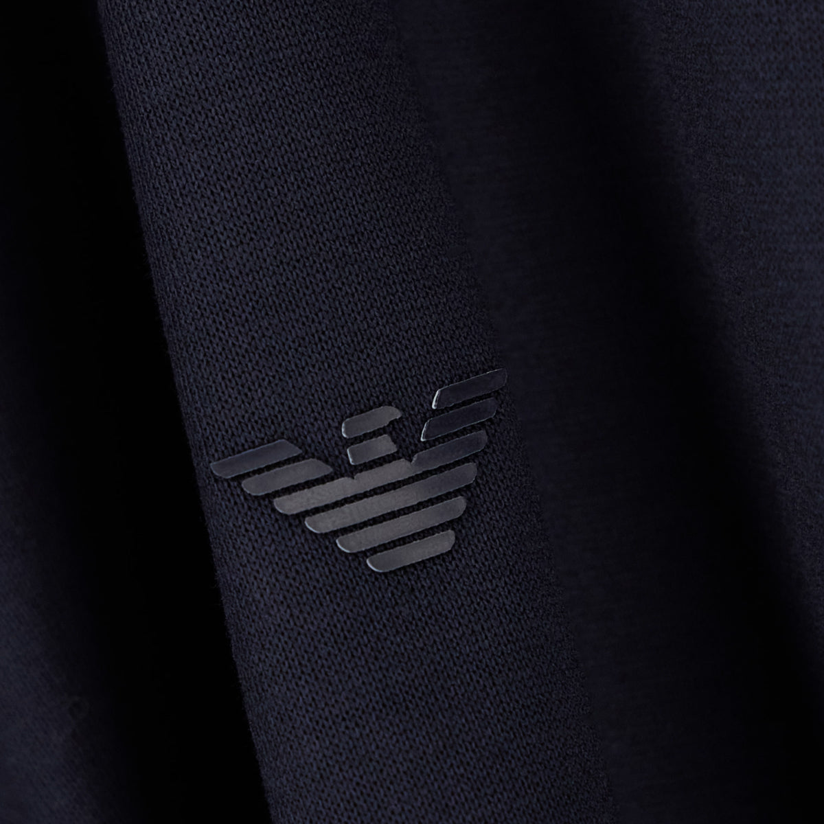Close-up of a dark navy knit fabric with a tonal black embroidered Emporio Armani eagle logo. Emporio Armani Polo available at Sakks Menswear in Southampton.