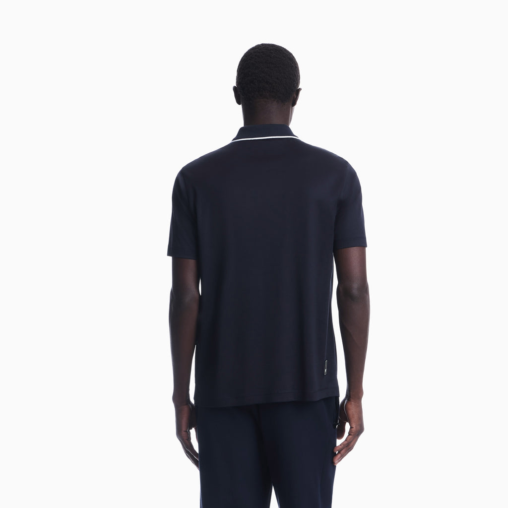 A man viewed from the back wearing a dark navy short-sleeve polo shirt with a thin white stripe detail on the collar, paired with matching navy trousers, against a solid white background. Emporio Armani Polo available at Sakks Menswear in Southampton.