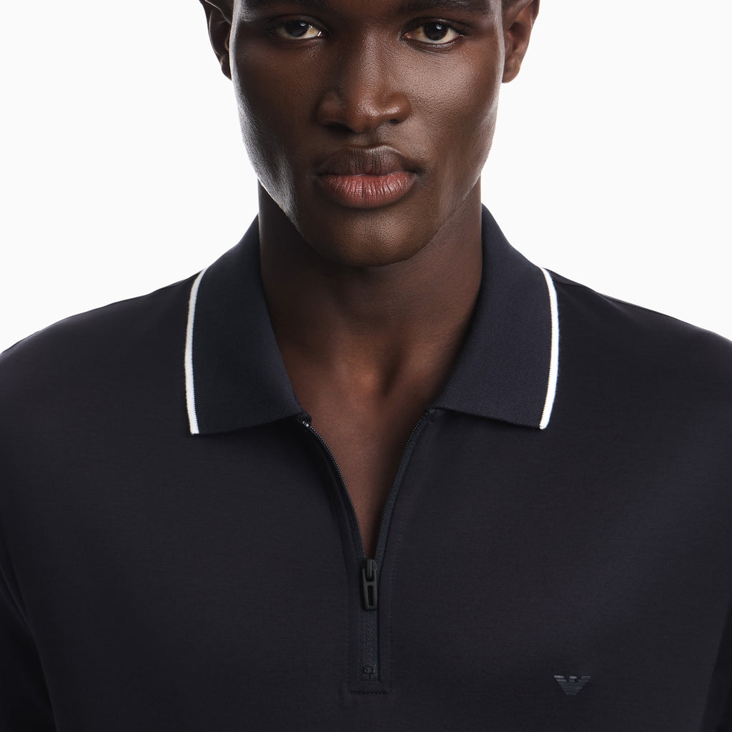 Close-up of a man wearing a dark navy polo shirt with a white contrast trim on the collar and a small embroidered logo on the chest, against a plain white background. Emporio Armani Polo available at Sakks Menswear in Southampton.