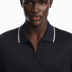 Close-up of a man wearing a dark navy polo shirt with a white contrast trim on the collar and a small embroidered logo on the chest, against a plain white background. Emporio Armani Polo available at Sakks Menswear in Southampton.