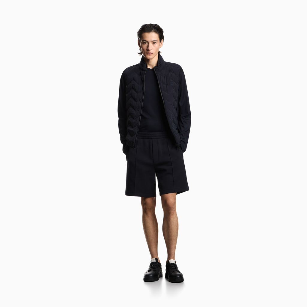 A male model stands against a plain white studio background wearing a dark navy quilted jacket over a matching dark navy top and shorts, with black sneakers. Emporio Armani Shorts available at Sakks Menswear in Southampton.