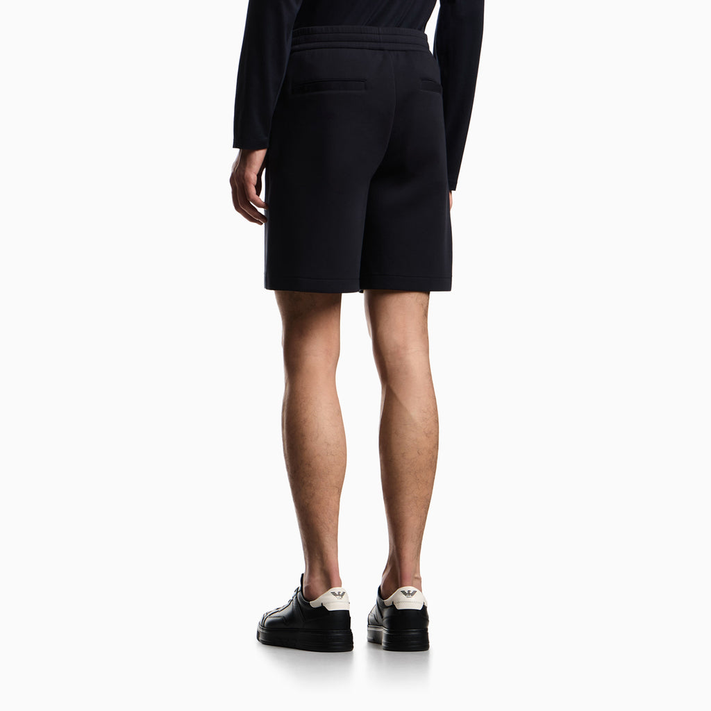 Rear view of a model wearing black knit shorts with a drawstring waist, a black long-sleeve top, and black leather sneakers with white heel tabs, against a plain white background. Emporio Armani Shorts available at Sakks Menswear in Southampton.