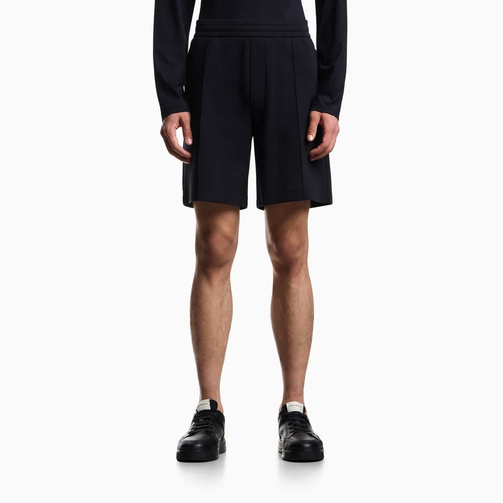 A model from the waist down wearing dark navy pleated athletic shorts, a matching long-sleeve top, and black sneakers against a light gray studio background. Emporio Armani Shorts available at Sakks Menswear in Southampton.