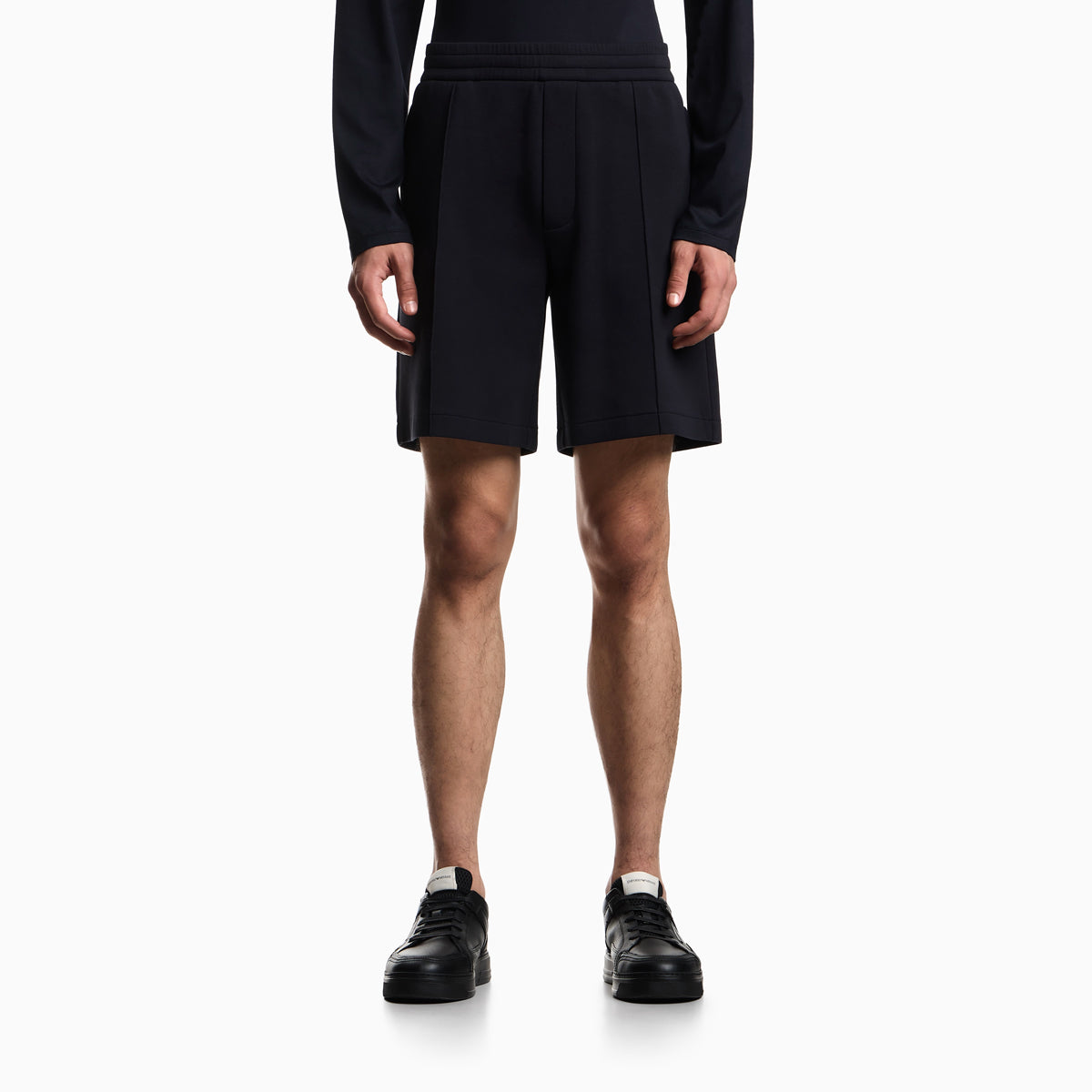 A model from the waist down wearing dark navy pleated athletic shorts, a matching long-sleeve top, and black sneakers against a light gray studio background. Emporio Armani Shorts available at Sakks Menswear in Southampton.