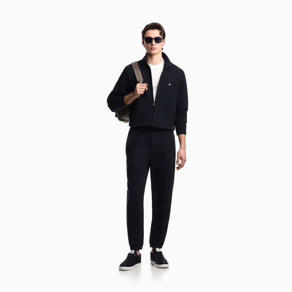 A man modeling a black zip-up jacket and matching black track pants, styled with a white t-shirt, black sneakers, sunglasses, and a gray shoulder bag against a plain white background. Emporio Armani Sweatshirt available at Sakks Menswear in Southampton.